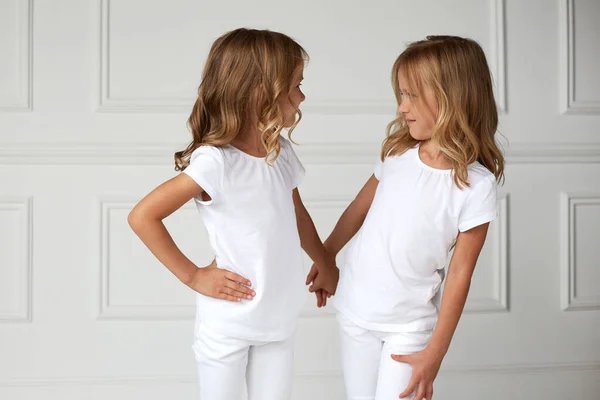 Twee kleine kinderen meisjes in witte kleren, over witte achtergrond kinderen studio portret. Tweeling meisjes kijken elkaar aan. — Stockfoto