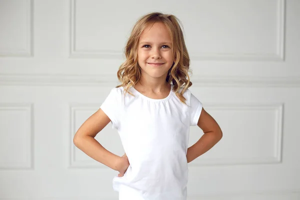 Voorportret van een klein meisje lachend en kijkend naar de camera, gekleed in een wit T-shirt, met haar handen op haar middel, op een witte achtergrond. — Stockfoto