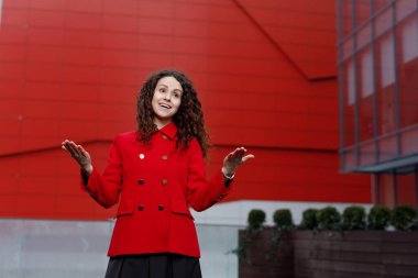 Kırmızı ceketli genç bir kadın, elleri kenarda duruyor, şaşkınlık içinde, bir binanın arka planını gösteriyor..