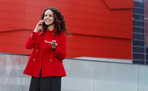 Kırmızı ceketli genç bir kadının güzel portresi, cep telefonuyla konuşuyor, dışarıda yürüyor, binaya karşı.. — Stok fotoğraf