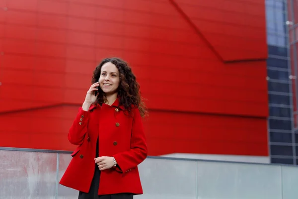 Porträt einer lächelnden brünetten, lockigen jungen Frau, trägt einen roten Anzug, telefoniert mit dem Handy, vor dem Hintergrund des Gebäudes. — Stockfoto