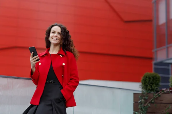 一个穿着红色衣服的优雅微笑的年轻女子的户外肖像，手里拿着智能手机，走在外面. — 图库照片