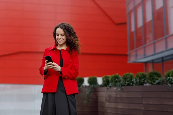 Neşeli genç esmer kadın, ceketini giyiyor, akıllı telefonundan konuşma okuyor, binanın arka planında.. — Stok fotoğraf