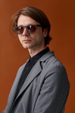 Kırmızı güneş gözlüklü çekici erkek model, sağlam bir duruşla duruyor, kahverengi stüdyo arka planında kameraya bakıyor. Dikey görünüm.