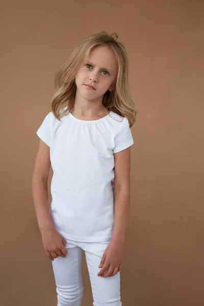 Verticaal beeld van een klein meisje met blond haar helemaal in het wit gekleed, met een fronsend gezicht, op een beige achtergrond. — Stockfoto