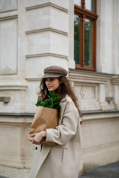 Portrét stylové mladé ženy kráčející v chladném dnu.s papírovou krabičkou zelených listů rostlin na městském pozadí. — Stock fotografie