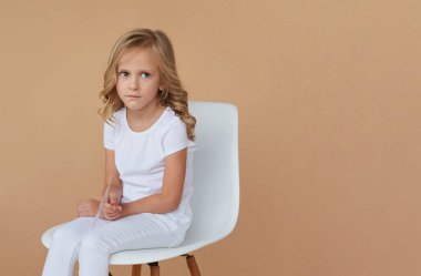 Closeup front portrait of pretty little girl with wavy blond hair, dressed in white clothes, sitting on chair, over beige background.