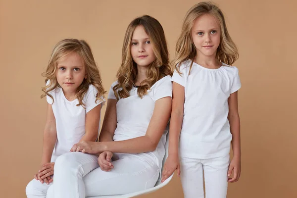 Voorzijde close-up portret van drie zussen met blond krullend haar gekleed in witte kleren, geïsoleerde beige achtergrond. — Stockfoto