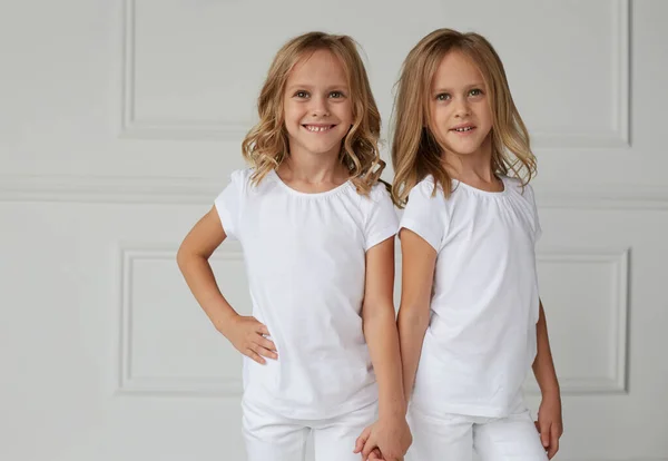Twee tweelingzussen in witte kleren die elkaars hand vasthouden op een witte achtergrond. Het concept van de levensstijl van kinderen. Horizontaal zicht. — Stockfoto