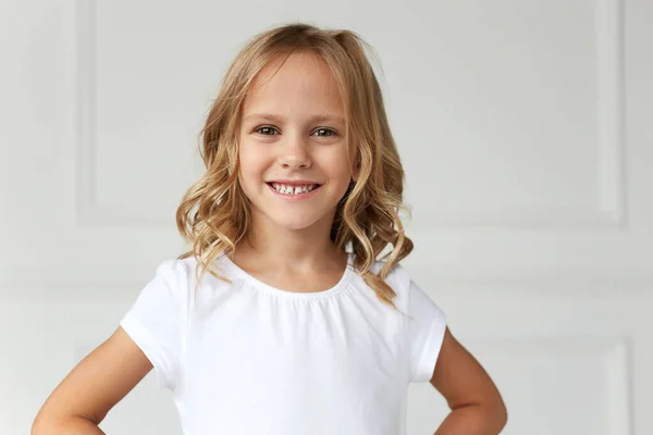 Voorportret van een klein meisje glimlachend en kijkend naar de camera, gekleed in een wit shirt, met haar handen op haar middel. moedig en mooi meisje, op een witte achtergrond. — Stockfoto