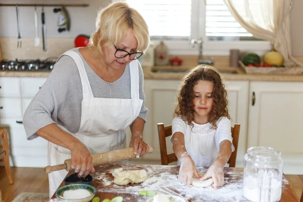 Grandma in white apron with cute curly granddaughter spend time in the kitchen. Grandmother teach the niece to prepare.