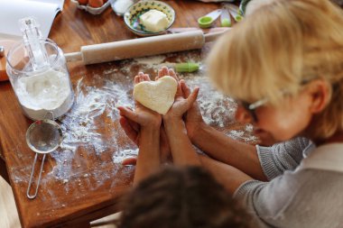 Frame image of a female and kid hands holding dough in heart shape top view. Baking ingredients on the dark table clipart