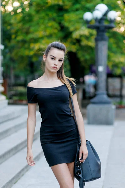 Fashion style portrait of young beautiful calm female model posing at city street in black slim dress and bag.
