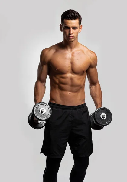 Front image of a confident young man shirtless torso portrait training with dumb-bell, isolated grey background. Vertical view. — Fotografia de Stock