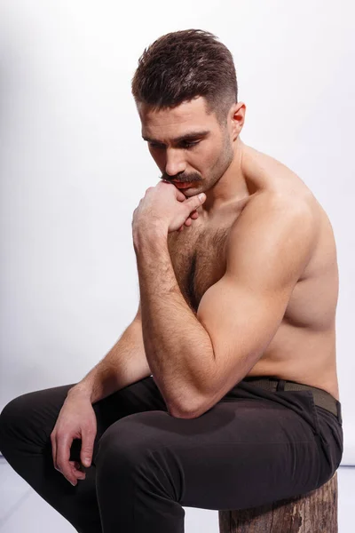 Homem atraente com bigode, sem camisa, mostrando bíceps e tríceps, sentado em um tronco de árvore, sobre fundo branco. — Fotografia de Stock