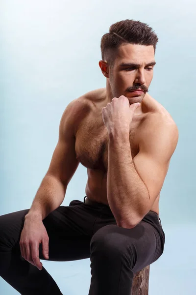 Young man with moustache, shirtless, showing biceps and triceps, seated on a tree trunk, over white background. — Stock Photo, Image