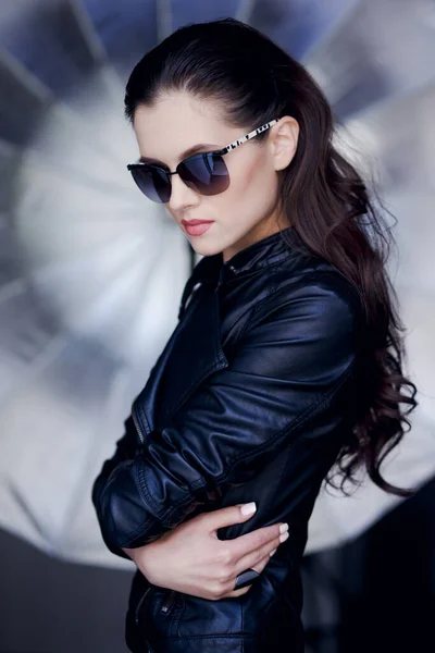 Selbstbewusste und sexy junge brünette Frau posiert in schwarzer Lederjacke und Sonnenbrille auf cremesilbernem Hintergrund. — Stockfoto