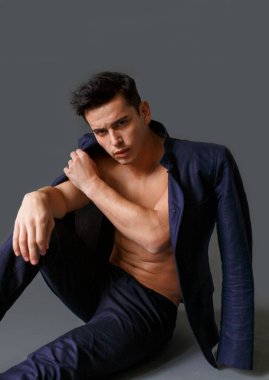 Yakışıklı, esmer, omuzlarında ceketi, çıplak gövdesi olan, gri arka planda oturan seksi bir adam..