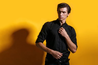 Siyah elbiseli, stil sahibi, sarı arka planda poz veren genç bir adamın yakın plan portresi..