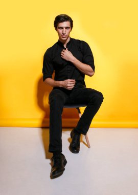 Sarı arka planda oturan, siyah elbiseli, neşeli, yakışıklı, güçlü genç bir adamın tam boy görüntüsü..