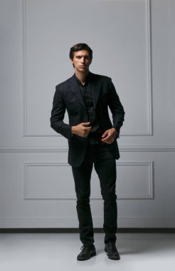 Şık, modaya uygun bir adamın kameraya bakarken giysisini ayarlarken, neoklasik duvarın yanındaki stüdyoda poz verirken tam boy dikey görüntüsü..