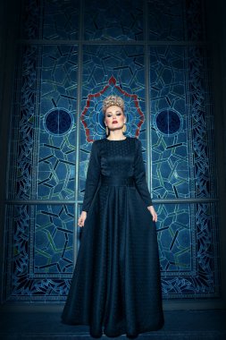 Uzun siyah bir elbise giymiş, zarif saç stili, harika bir taç ve küpeleri olan sarışın bir mankenin tam boy görüntüsü..