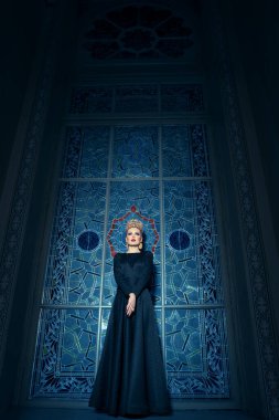 Vertical view. Full length image of a great young blonde female model, dressed in a long black dress, with elegant hairstyle crown and earrings.