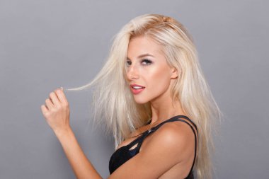 Beauty profile portrait. Hot blonde girl in black bra, on grey background, arranges her hair, looking at camera.