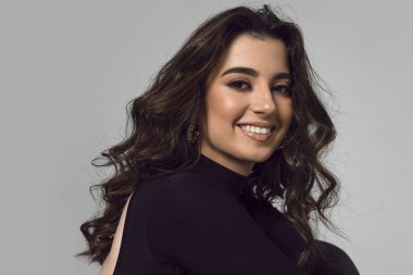 Closeup front portrait of a smiling young woman, model with brunette curly hairstyle and make up, looking at camera, over grey background.