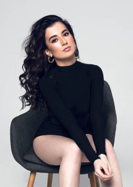 Front view of a sensual young woman with hairstyle wear black bodysuit sitting on chair, looking at camera, on a grey background. Vertical view.
