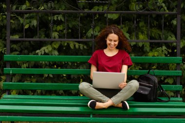 Dizüstü bilgisayarla gülümseyen, parkta bankta oturan bir kadının portresi..