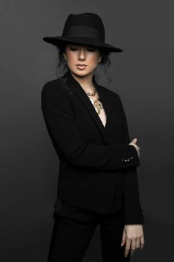 Front portrait of young splendid brunette woman in black suit and hat with makeup, with closed eyes, over grey background.