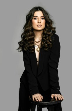 Güzel, genç, esmer, kıvırcık saçlı, gece makyajlı, siyah takım elbiseli, kameraya bakan, gri arka planlı bir kadın..