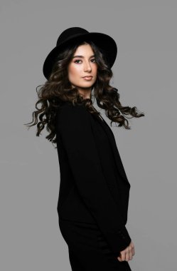 Siyah takım elbiseli, makyajlı, kıvırcık saçlı, gri arka planda harika bir esmer kadın..