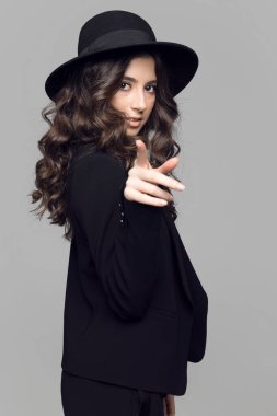 Güzel, genç, esmer, kıvırcık saçlı, gece makyajlı, siyah takım elbiseli, kameraya bakan, gri arka planlı bir kadın..