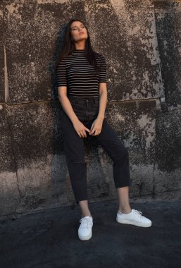 Kahverengi, uzun saçlı güzel bir kadın, eski siyah bir duvara yaslanmış kameraya bakıyor..