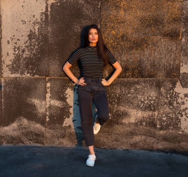 Kahverengi, uzun saçlı güzel bir kadının tam boy görüntüsü eski siyah bir duvara yaslanmış kameraya bakıyor..