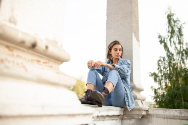 Portrait of a single pensive teenage girl dressed in denim and boots, sitting, looking down.