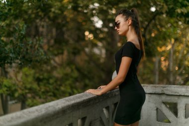 Siyah ince elbiseli, güneş gözlüklü, saçları at kuyruklu, balkonda kalan şehvetli genç bir kadının profili..