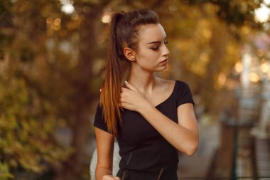 Siyah ince elbiseli, saçları at kuyruklu, balkonda poz veren, ılık ışıklı, bulanık ağaç arka planlı esmer bir kadın..