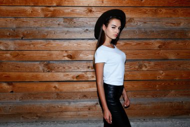 Şapkalı bir kızın profil portresi, düz saçları, deri pantolon ve tahta duvarın yanındaki tişört..