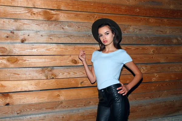 Şapkalı bekar bir kadın mankenin portresi, düz saçı, deri pantolon ve mavi t-shirt 'ü, tahta duvarın yanında, geceleri yürüyor..