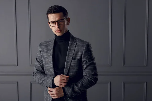 Um homem bonito em uma jaqueta cinza e gola alta preta, de pé na frente e olhando confiante, no fundo da parede cinza. — Fotografia de Stock