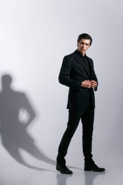 Genç adam stüdyoda tavır takınıyor, kameraya bakıyor, takım elbisesini ayarlıyor, beyaz arka planda izole edilmiş..