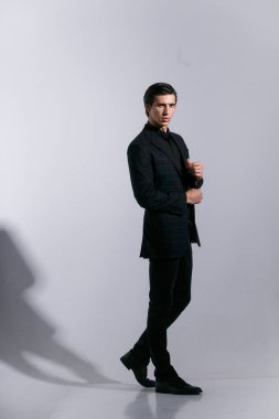 Zarif takım elbiseli bir adam, beyaz arka planda izole bir tavırla poz veriyor. Ceketinin düğmesine basıyor. Dikey görünüm.