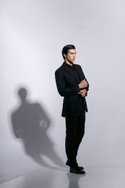 Takım elbiseli bir adamın uzun bir portresi, beyaz arka planda tavır takınarak poz veriyor. Ceketinin düğmesine basıyor..