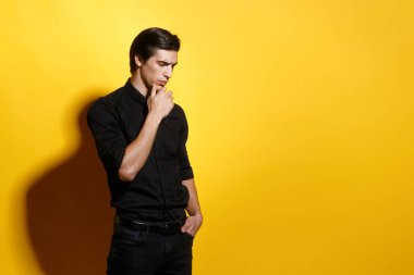 Siyah gömlekli ve pantolonlu şık bir erkek modelin profil portresi, elini cebine atıyor, kameraya bakıyor, sarı arka planda izole edilmiş, boşluğu kopyalıyor..