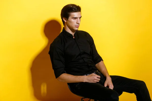 黒いシャツとパンツのスタイリッシュな男性モデルのプロフィール 椅子に座って真剣に 黄色の背景に孤立して見える コピースペース — ストック写真