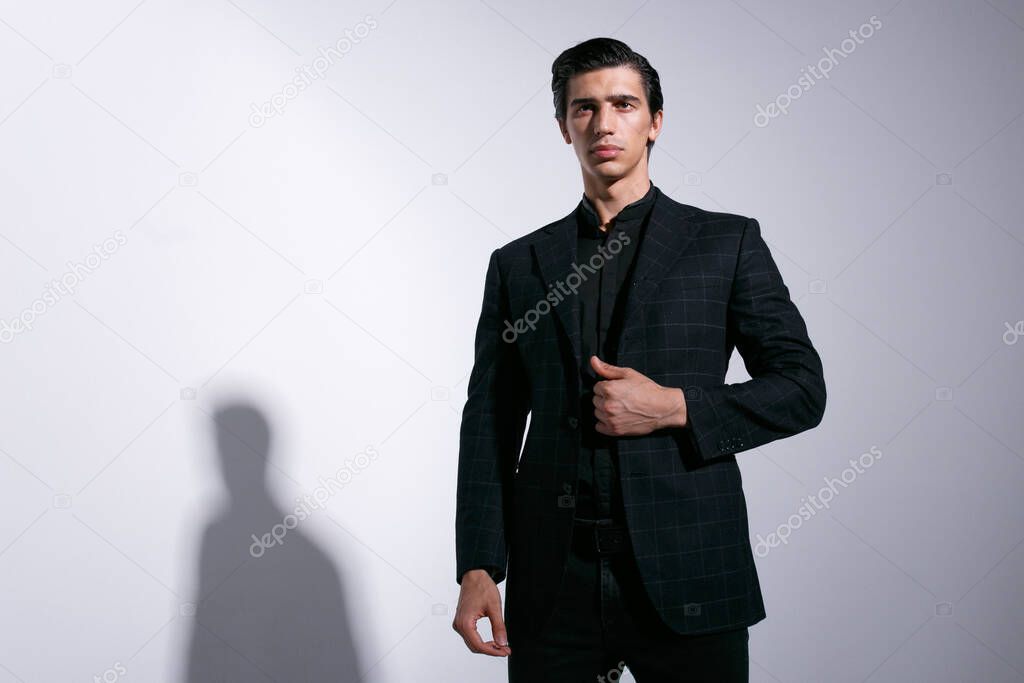 Young sexy handsome male model dressed in black suits on white studio background with shade on the wall. Horizontal indoors shot.