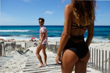 Gülümseyen çekici bir gencin arka görüntüsü, kumsaldaki seksi genç bayanla tanışmak için geri döner..